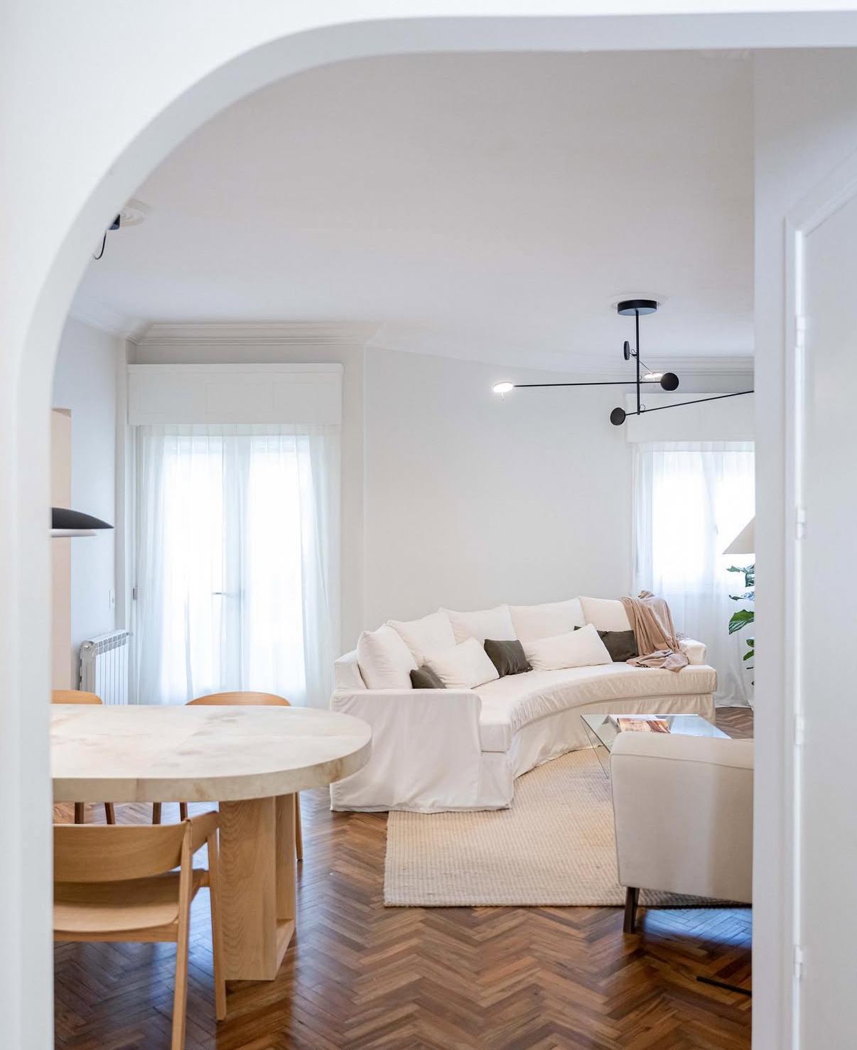 Living luminoso con sofá curvo blanco, piso de parquet espigado y comedor de madera clara visto desde un arco