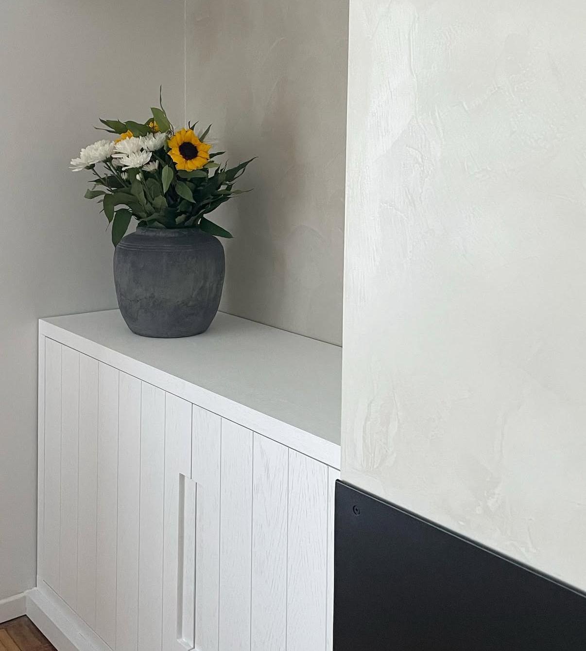 Mueble bajo de madera lacada blanca con vasija gris y ramo de girasoles sobre pared de microcemento